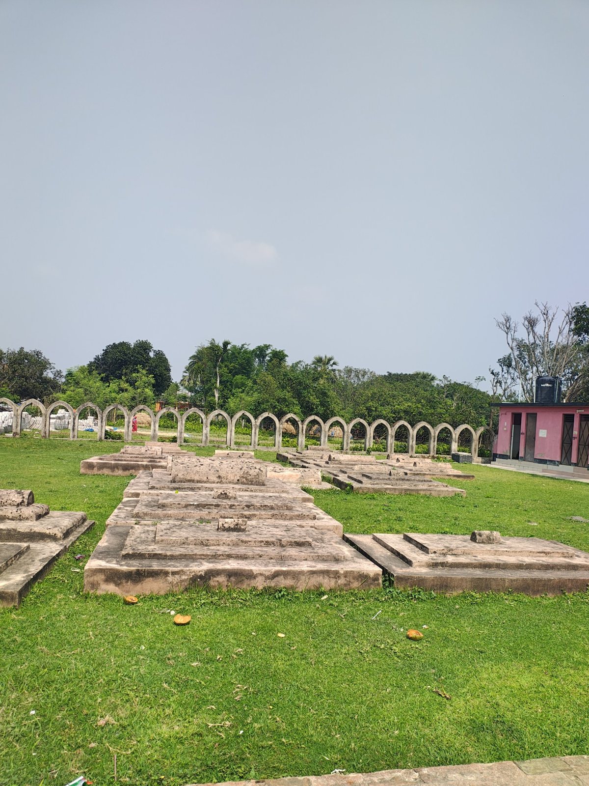 Stepped grave platforms and an arched heritage boundary wall among green trees in Chapainawabganj