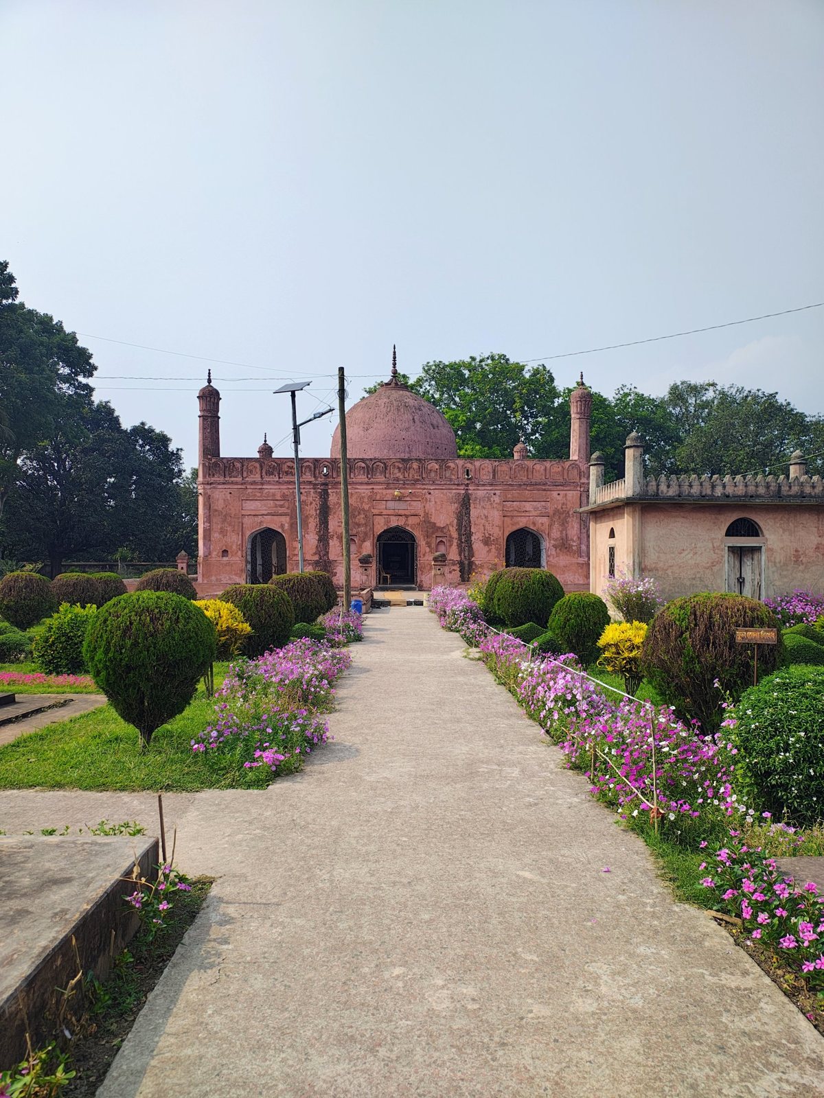 Pink-toned domed heritage-style building at the end of a flower-lined garden path in Chapainawabganj