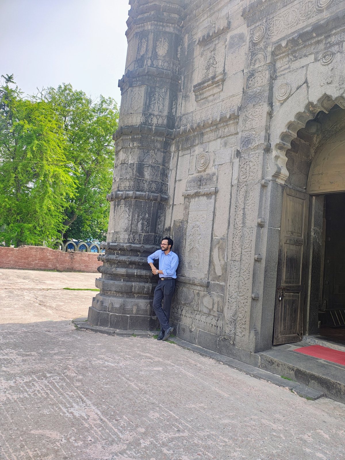 Sayedul Abrar standing by the carved pillar near Sona Masjid's arched entrance, with green trees behind